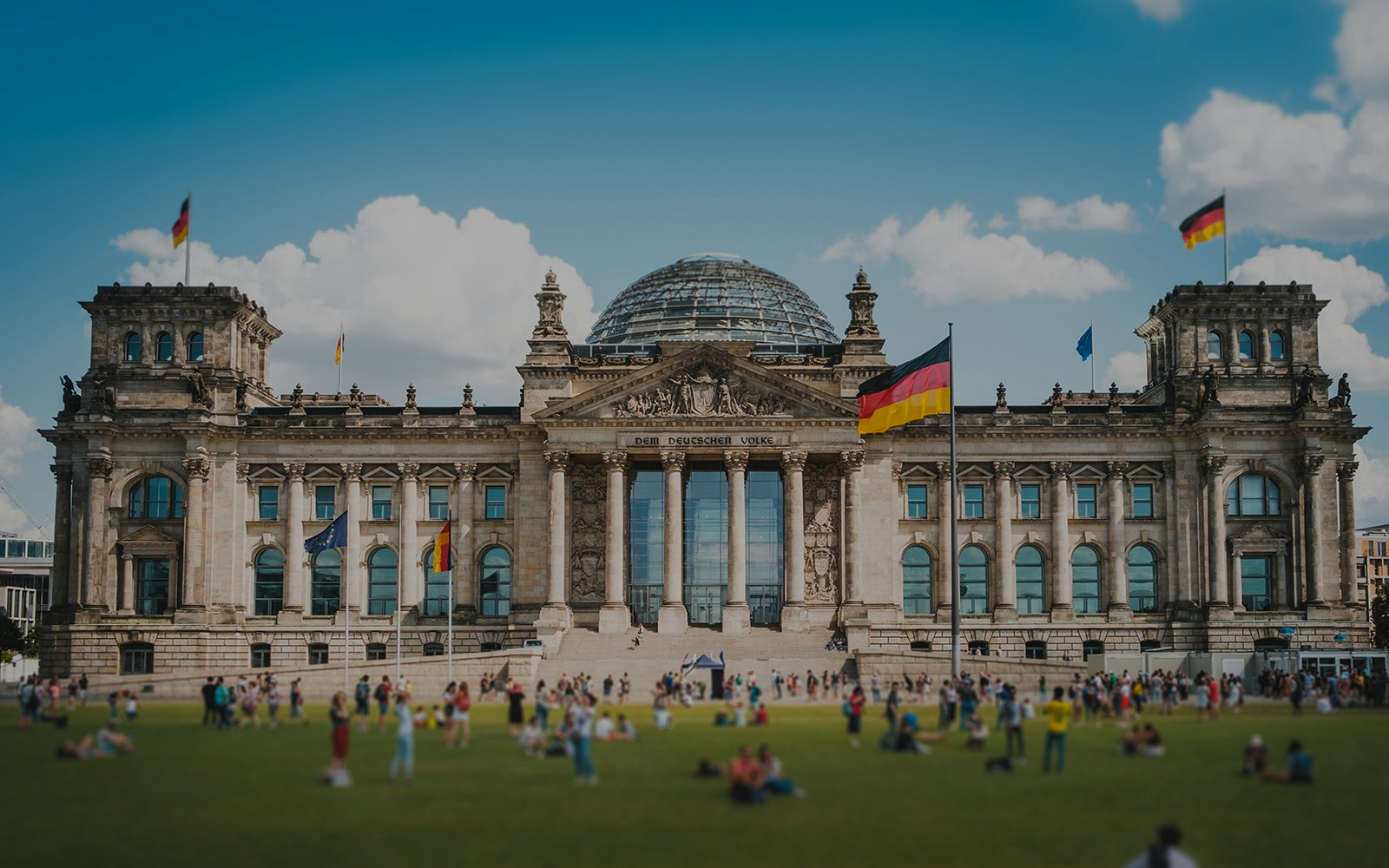 Reichstag building in Berlin with visitors on the lawn, German flags flying.