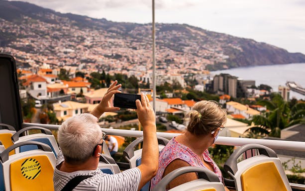 Tourists on Madeira hop-on hop-off bus capturing cityscape views.