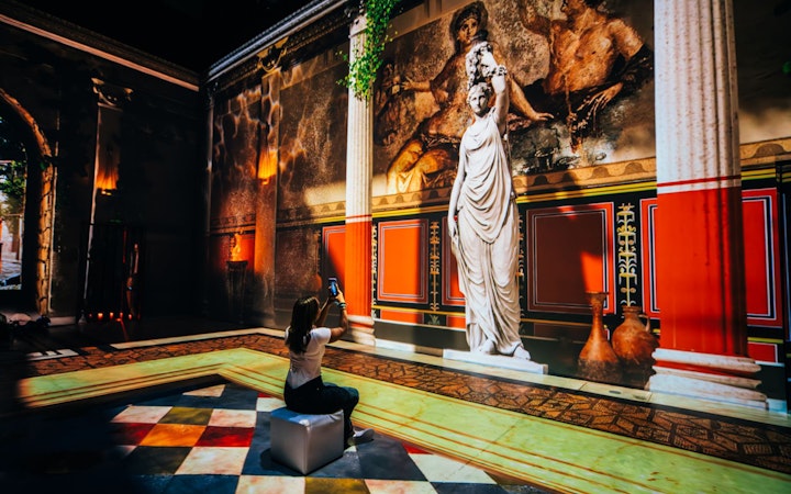 Interactive exhibit of ancient Pompeii with statue and fresco, visitor taking photo.