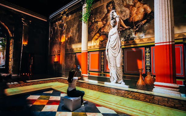 Interactive exhibit of ancient Pompeii with statue and fresco, visitor taking photo.