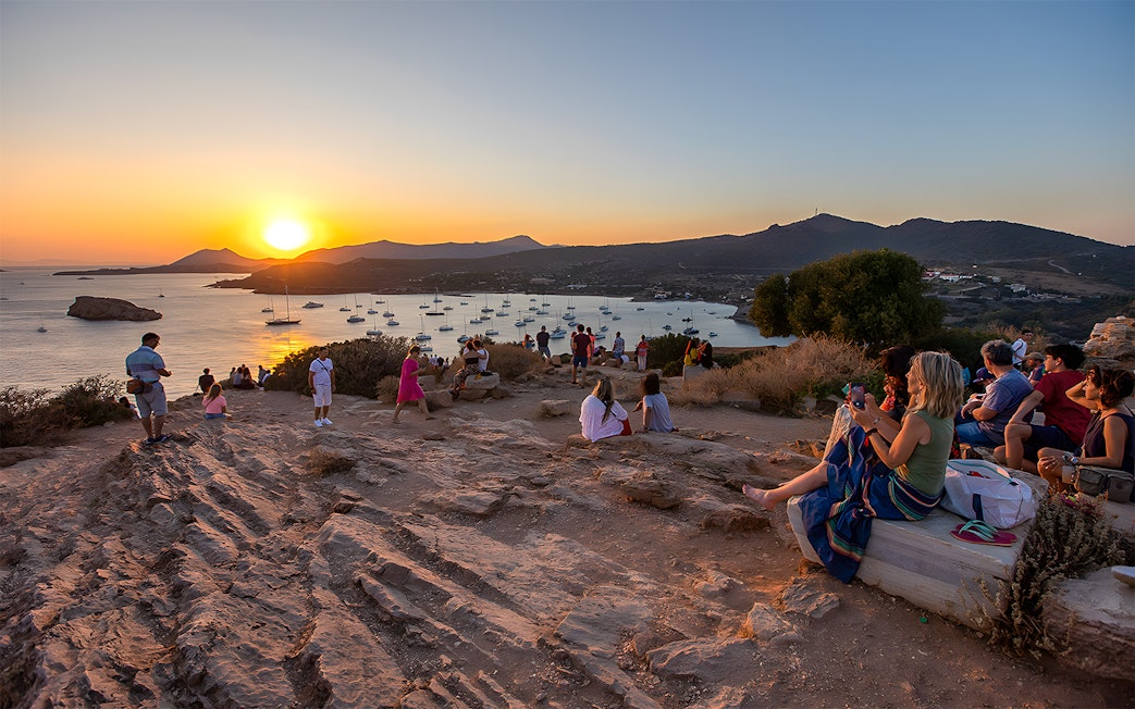 Tourists watching sunset at Archaeological Site of Sounion with sea and boats in view.