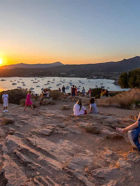 Tourists watching sunset at Archaeological Site of Sounion with sea and boats in view.