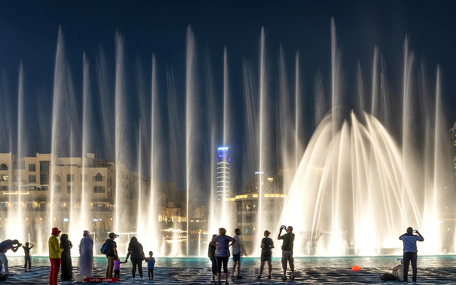 Dubai Fountain Boardwalk with tourists watching the fountain show at night, Burj Khalifa in the background.