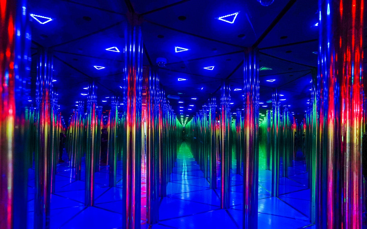 Tourists exploring colorful mirror maze with neon lights.