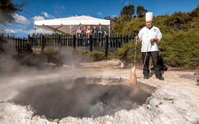 Chef demonstrating geothermal cooking at Te Puia, Rotorua, with tourists observing.