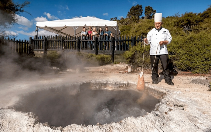 Chef demonstrating geothermal cooking at Te Puia, Rotorua, with tourists observing.