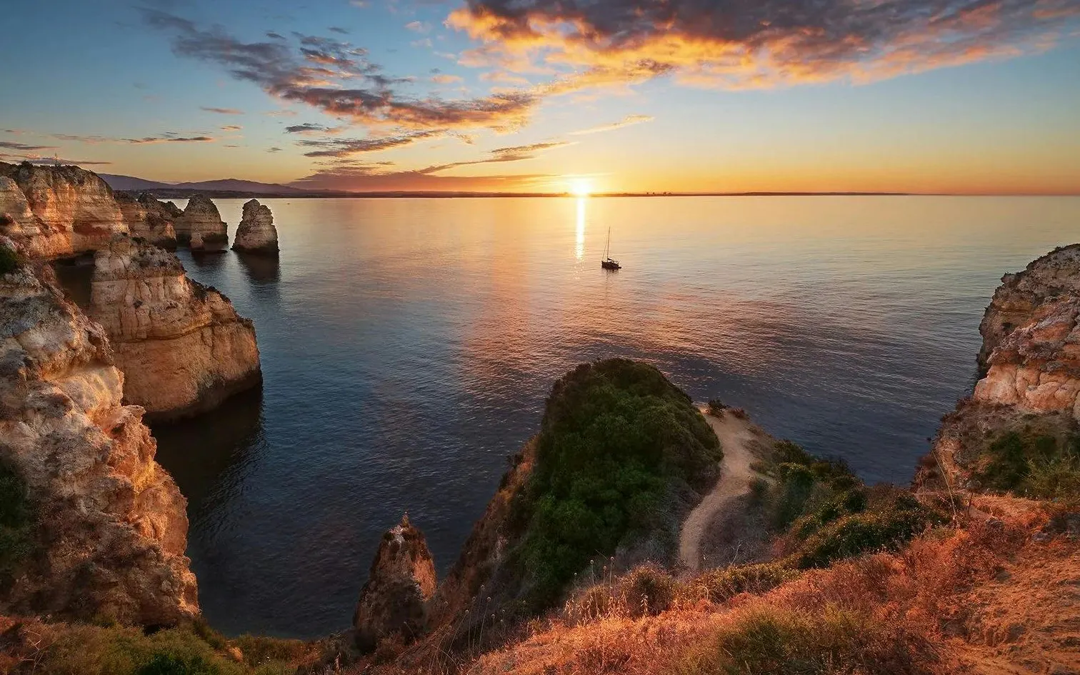 Cliffs overlooking the ocean at sunset in Algarve, Portugal with a sailboat in the distance.