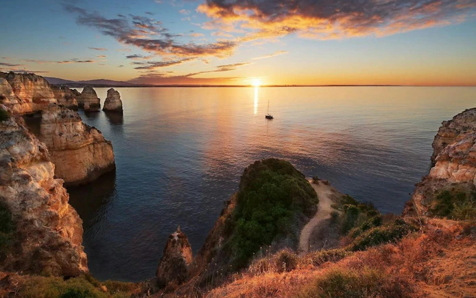 Cliffs overlooking the ocean at sunset in Algarve, Portugal with a sailboat in the distance.