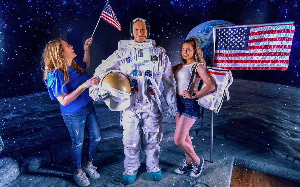 Neil Armstrong wax statue with visitors at Madame Tussauds, featuring lunar backdrop and American flag.