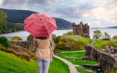 Tourist with Scottish umbrella views Urquhart Castle at Loch Ness, Scotland in autumn.