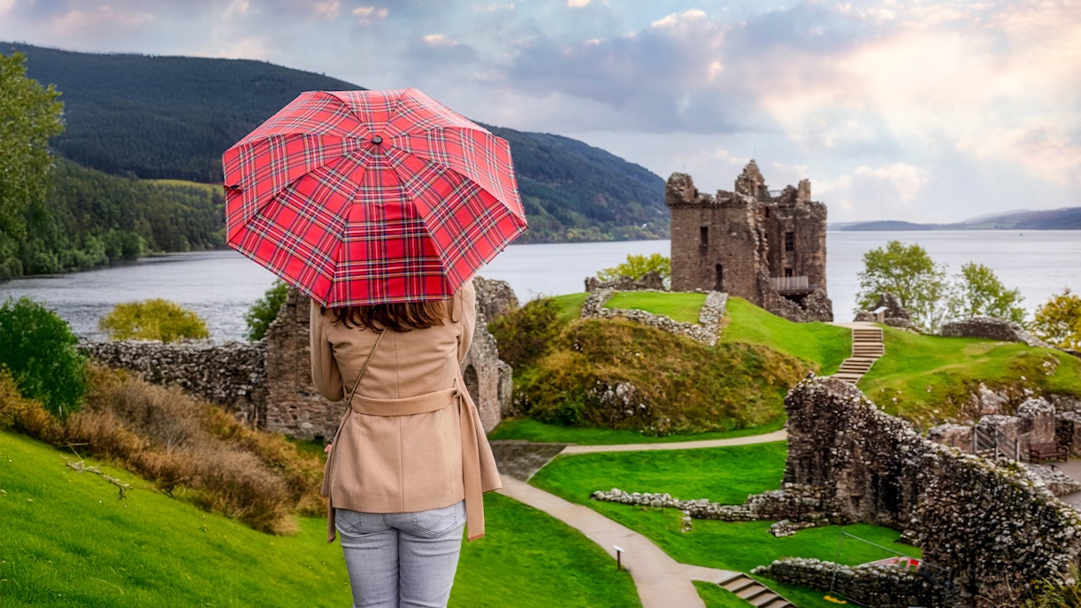 Tourist with Scottish umbrella views Urquhart Castle at Loch Ness, Scotland in autumn.