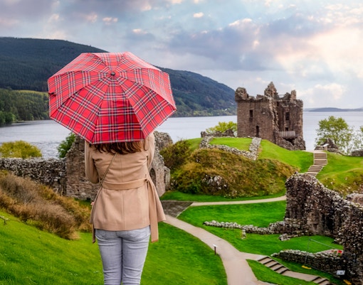 Tourist with Scottish umbrella views Urquhart Castle at Loch Ness, Scotland in autumn.