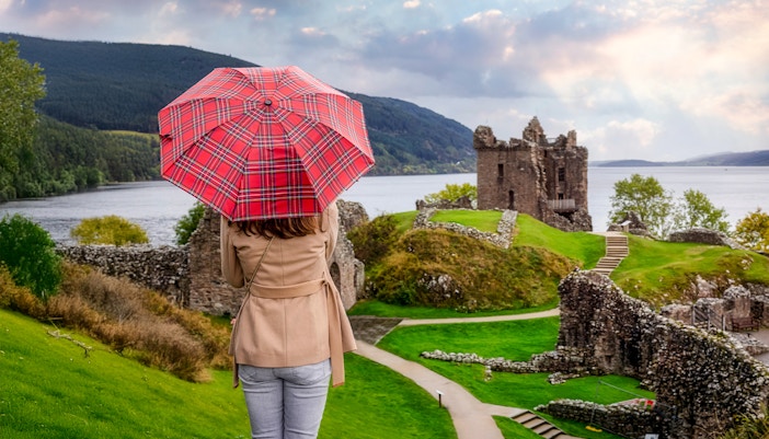 Tourist with Scottish umbrella views Urquhart Castle at Loch Ness, Scotland in autumn.