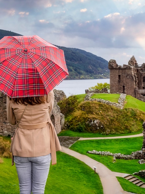 Tourist with Scottish umbrella views Urquhart Castle at Loch Ness, Scotland in autumn.