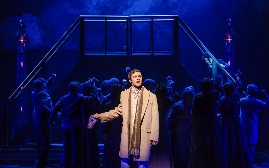 Performer in a coat on stage during Ragtime musical scene with ensemble in background.