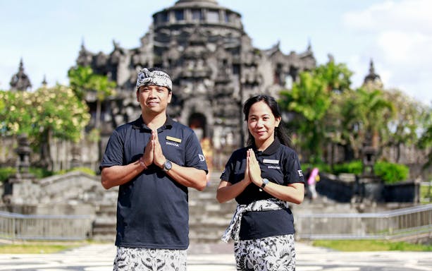 Tour guides in traditional attire at a Balinese temple, promoting airport transfers to Ngurah Rai.