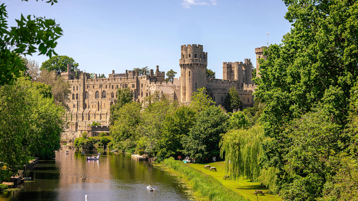 Warwick Castle by the river surrounded by lush greenery, showcasing medieval architecture.