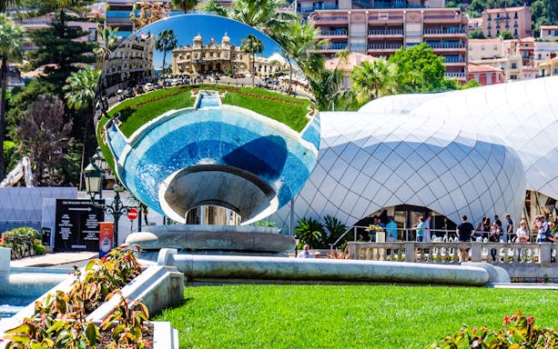 Reflection of Monte Carlo Casino in a large mirrored sculpture, surrounded by gardens.