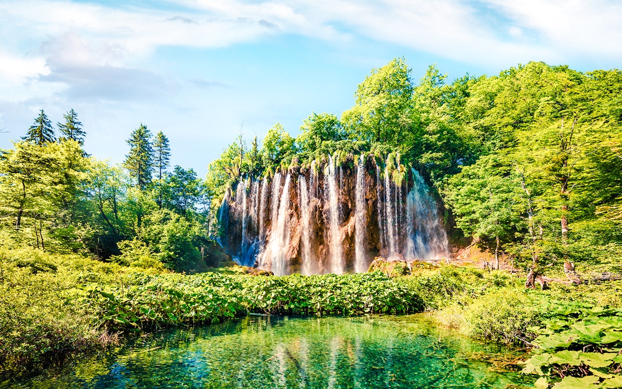 Waterfall cascading into a clear pool surrounded by lush greenery in Plitvice Lakes National Park.