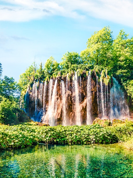 Waterfall cascading into a clear pool surrounded by lush greenery in Plitvice Lakes National Park.