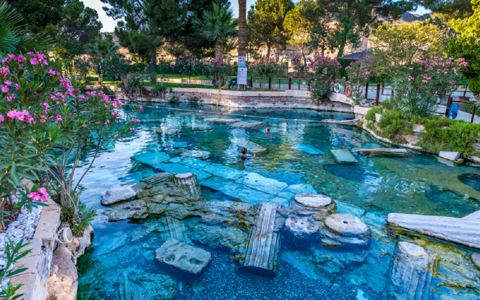 Ancient thermal pool with submerged ruins at Pamukkale, Turkey, surrounded by lush greenery.