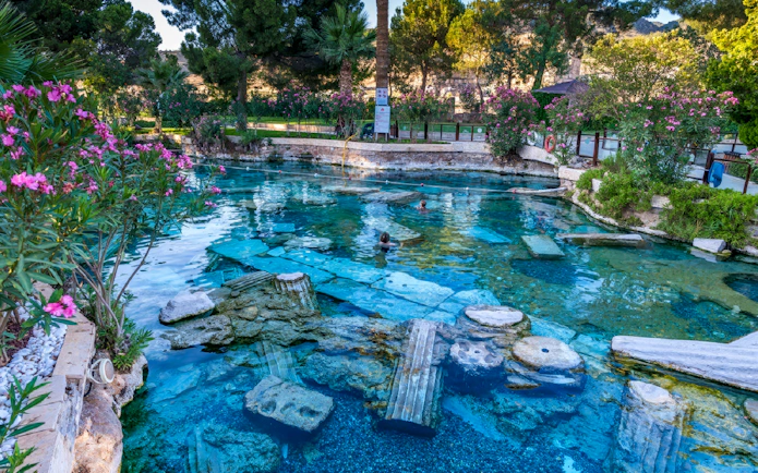 Ancient thermal pool with submerged ruins at Pamukkale, Turkey, surrounded by lush greenery.