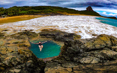 Person relaxing in Buraco do Galego natural pool, Noronha, with ocean and hills in background.