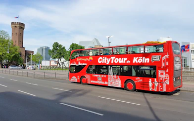 Red double-decker Hop on Hop off tour bus in Cologne near historic tower.