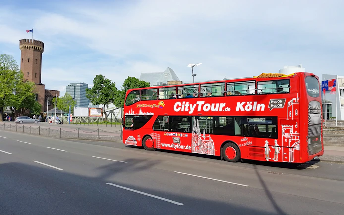 Red double-decker Hop on Hop off tour bus in Cologne near historic tower.