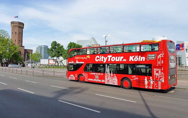 Red double-decker Hop on Hop off tour bus in Cologne near historic tower.