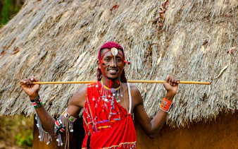 Maasai man in traditional attire holding a stick in front of a thatched hut, Zanzibar.