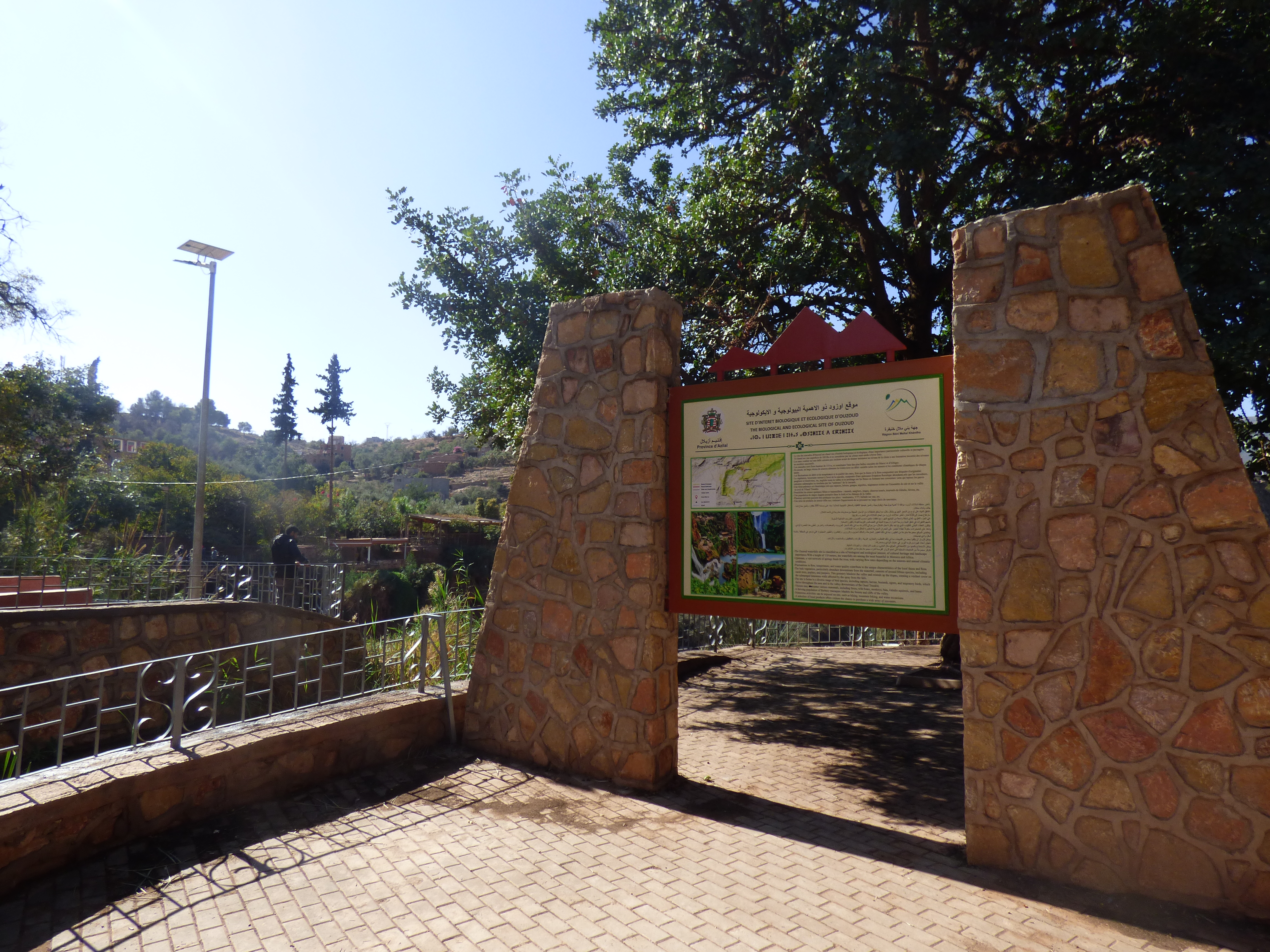 Entrance sign at Ouzoud Waterfalls trailhead, Morocco, surrounded by trees and stone structures.
