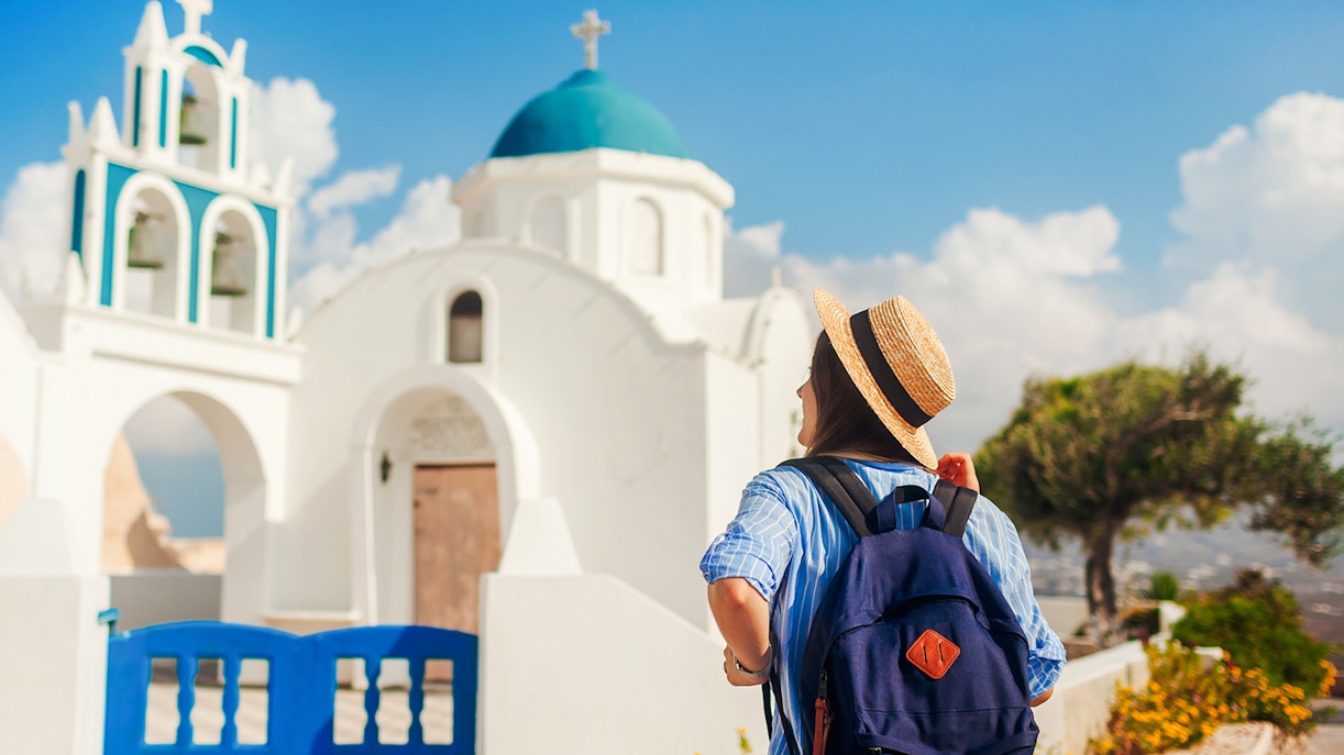 Tourist with backpack walking by a white church with blue dome in Santorini.