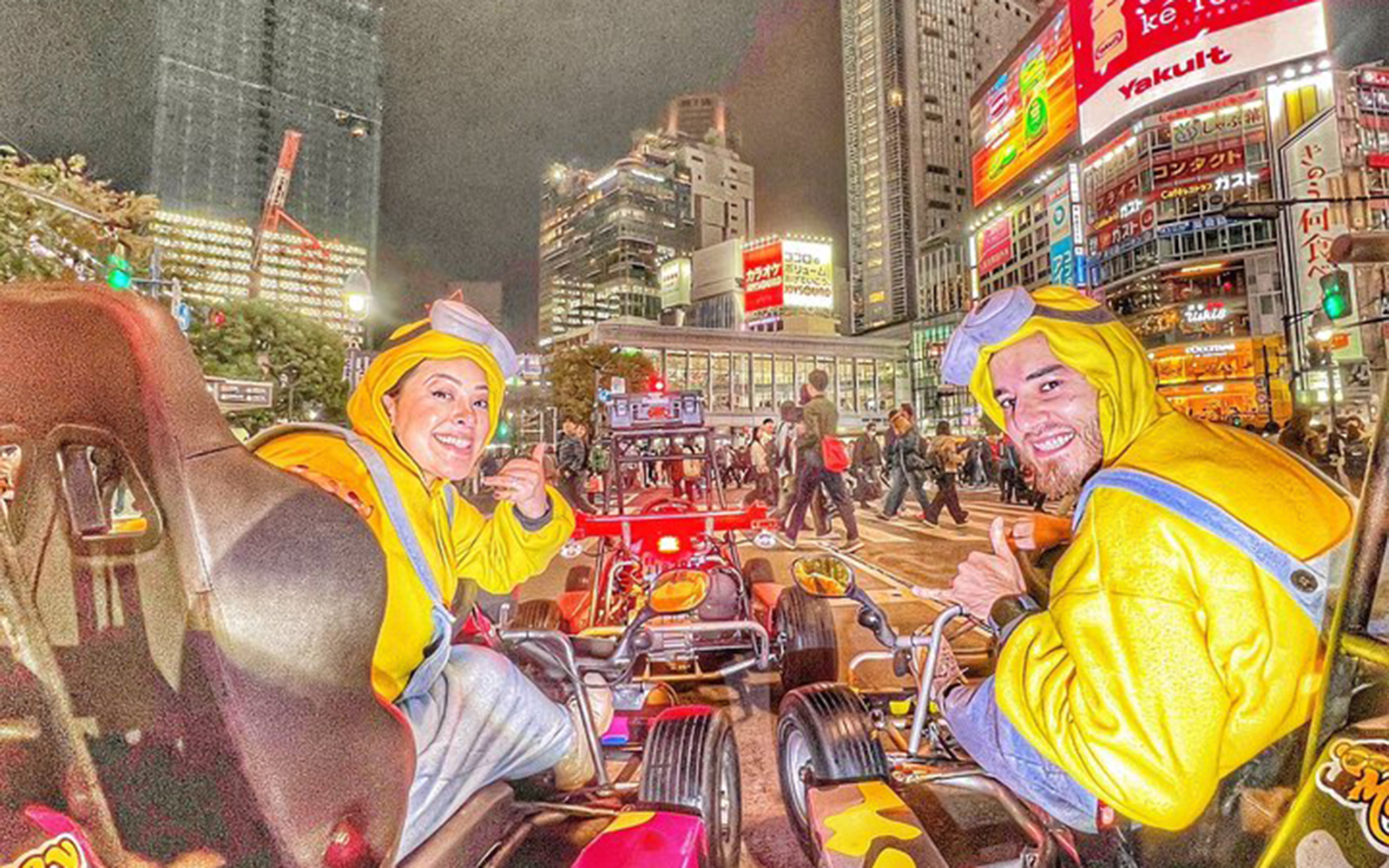 Two people in Minion costumes give thumbs-ups from go-karts on a busy street in Shibuya at night.