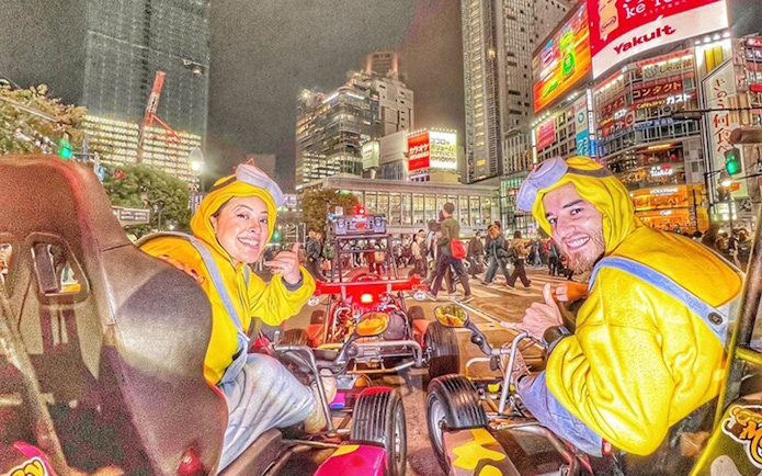 Two people in Minion costumes give thumbs-ups from go-karts on a busy street in Shibuya at night.