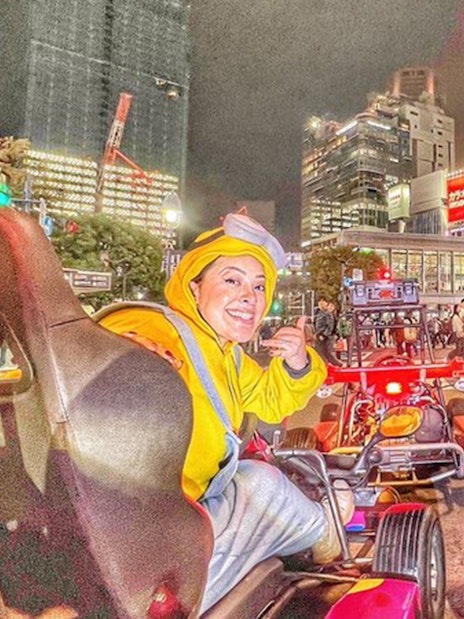 Two people in Minion costumes give thumbs-ups from go-karts on a busy street in Shibuya at night.