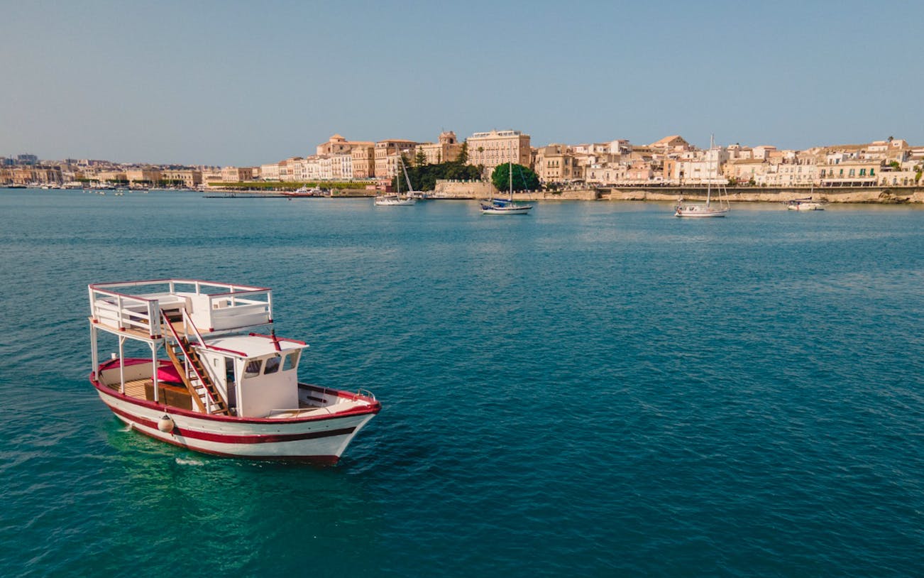 Boat on turquoise water near Ortigia Island, with historic buildings in the background.