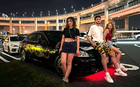 Group posing with custom cars at Daikoku PA during Tokyo Drift JDM Culture Tour.