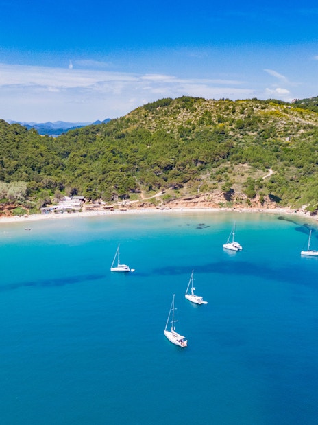 Sailboats anchored in the turquoise waters of Sunj Beach, surrounded by lush green hills.