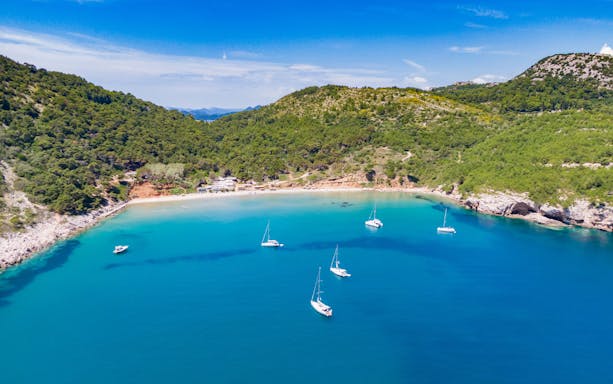 Sailboats anchored in the turquoise waters of Sunj Beach, surrounded by lush green hills.