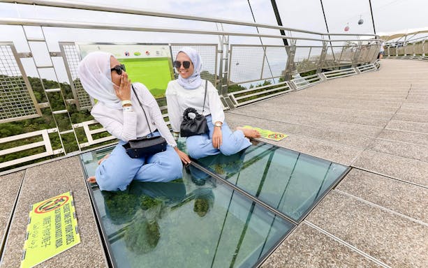 Visitors sitting on Langkawi SkyBridge glass floor with cable car in background.