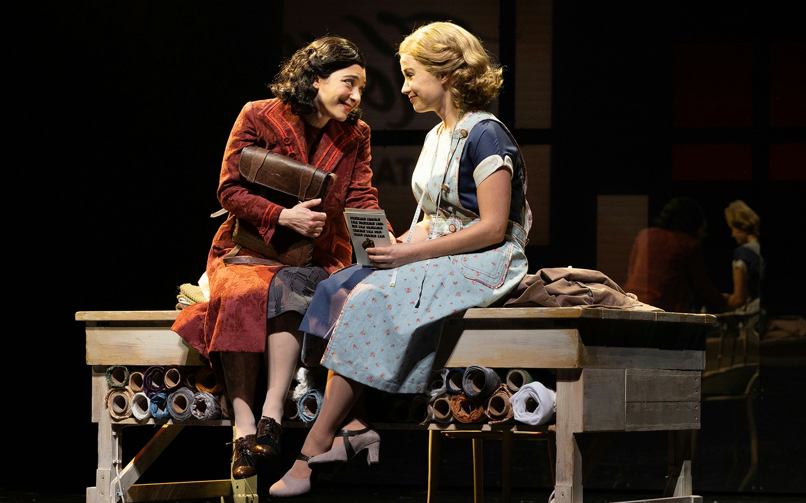 Two actors in period costumes sitting on a table, performing a scene from "Harmony: A New Musical.