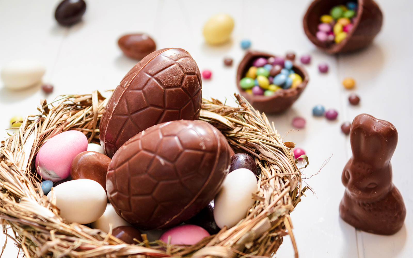 Chocolate eggs and Easter almonds in nest with chocolate bunny on white wooden table.