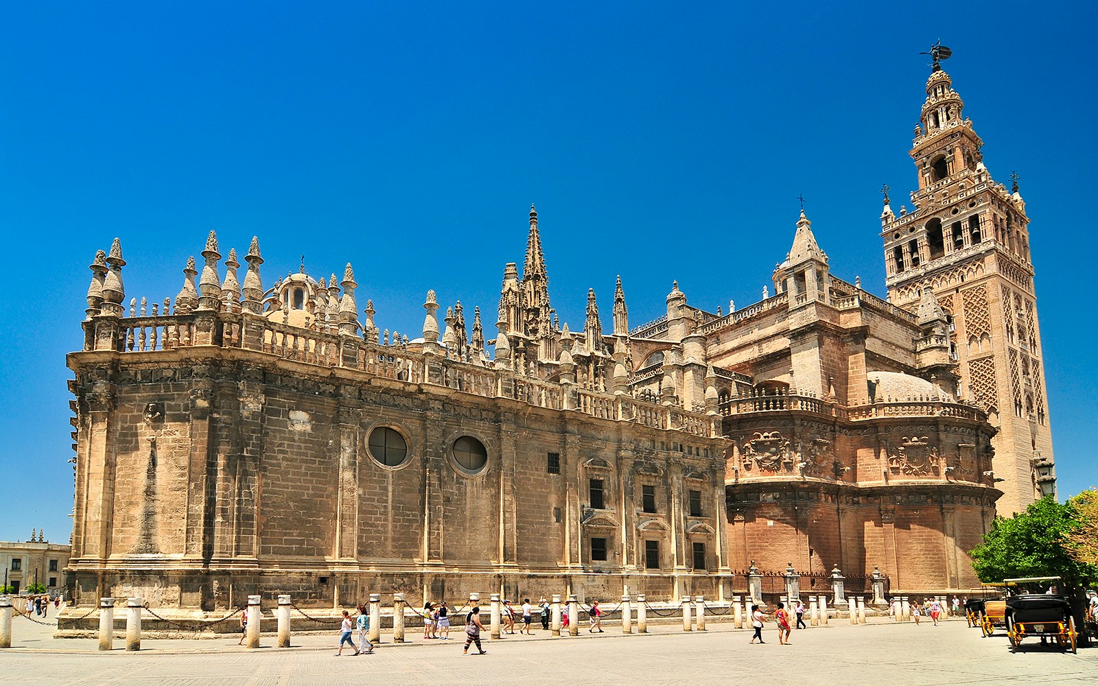 Seville Cathedral and Giralda tower under a clear blue sky, with tourists exploring the plaza.