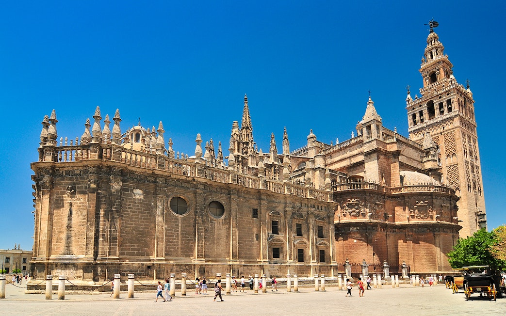 Seville Cathedral and Giralda tower under a clear blue sky, with tourists exploring the plaza.