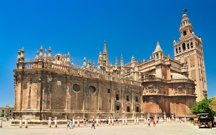 Seville Cathedral and Giralda tower under a clear blue sky, with tourists exploring the plaza.