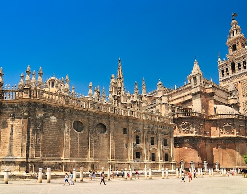 Seville Cathedral and Giralda tower under a clear blue sky, with tourists exploring the plaza.