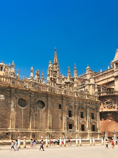 Seville Cathedral and Giralda tower under a clear blue sky, with tourists exploring the plaza.