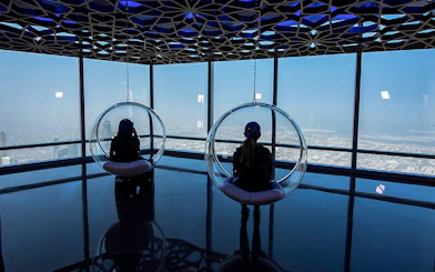 Visitors in glass chairs overlooking Dubai from Burj Khalifa observation deck.
