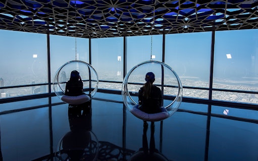 Visitors in glass chairs overlooking Dubai from Burj Khalifa observation deck.
